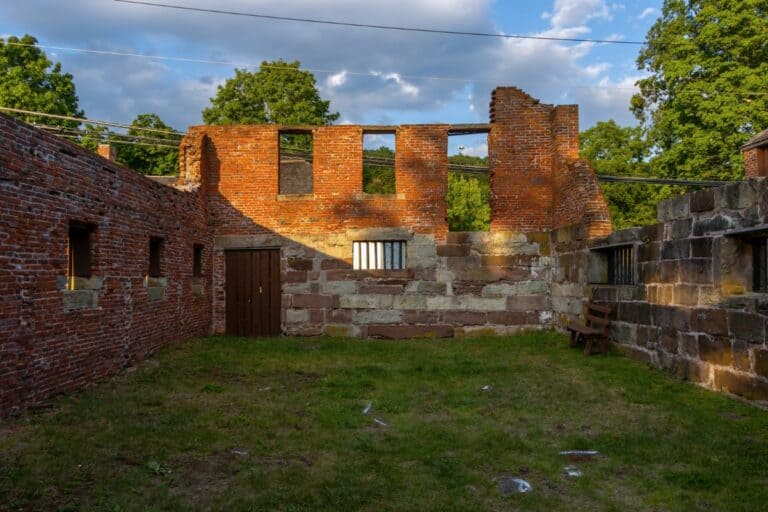Ruins of a brick and stone building with open roof, partial walls, and a wooden door, surrounded by grass and trees under a partly cloudy sky.