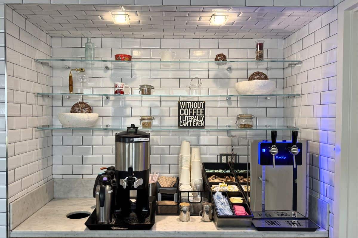Coffee station with a coffee maker, espresso machine, cups, condiments, and shelves holding jars, bowls, and a sign reading “Without coffee I literally can’t even.”.