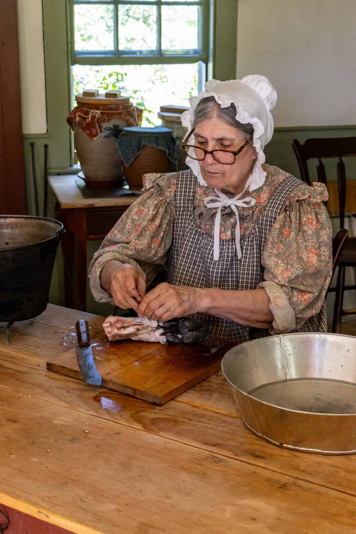 An older woman in period clothing prepares meat at a wooden table in a historic kitchen, with a knife, a metal basin, and crockery visible nearby.