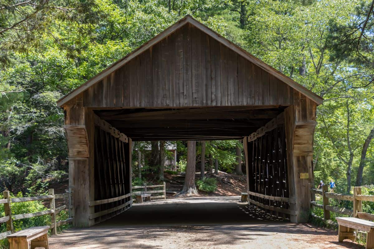 A wooden covered bridge with lattice sides crosses a path surrounded by trees and wooden fences on a sunny day.