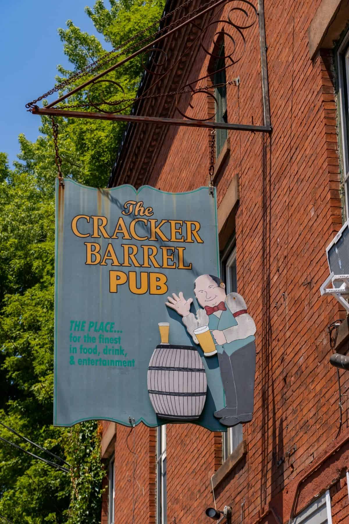 Sign for The Cracker Barrel Pub featuring an illustration of a man with beer mugs by a barrel, hanging on a brick building.