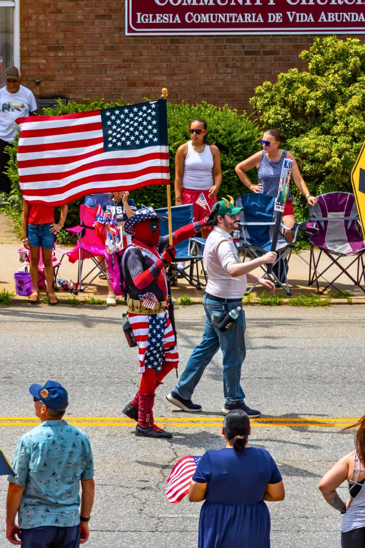 Two people walk in a parade; one is dressed in a superhero costume holding a large U.S. flag, while others watch from the sidewalk with chairs and flags.