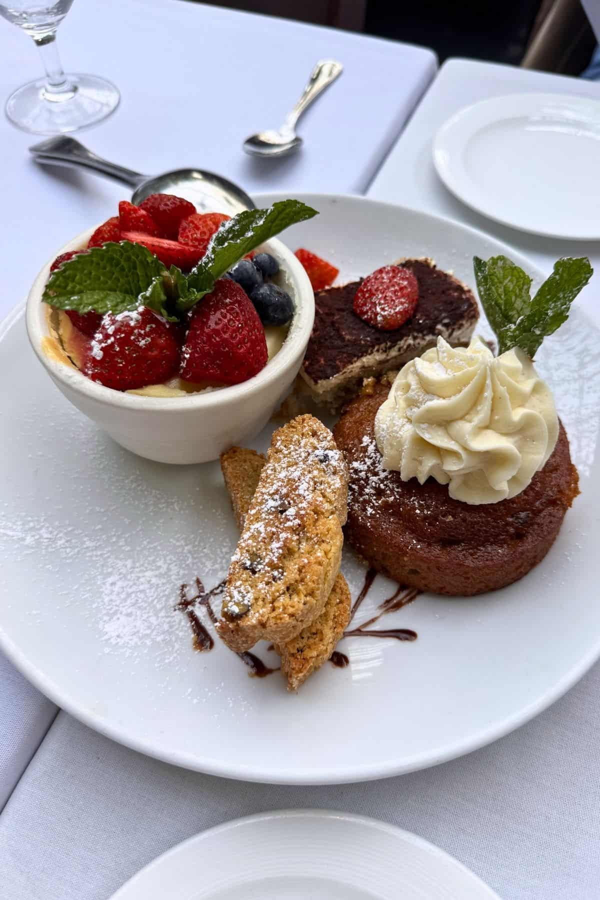 A white plate with tiramisu, a cup of berries on cream, biscotti, and a cake topped with whipped cream and mint, all dusted with powdered sugar.