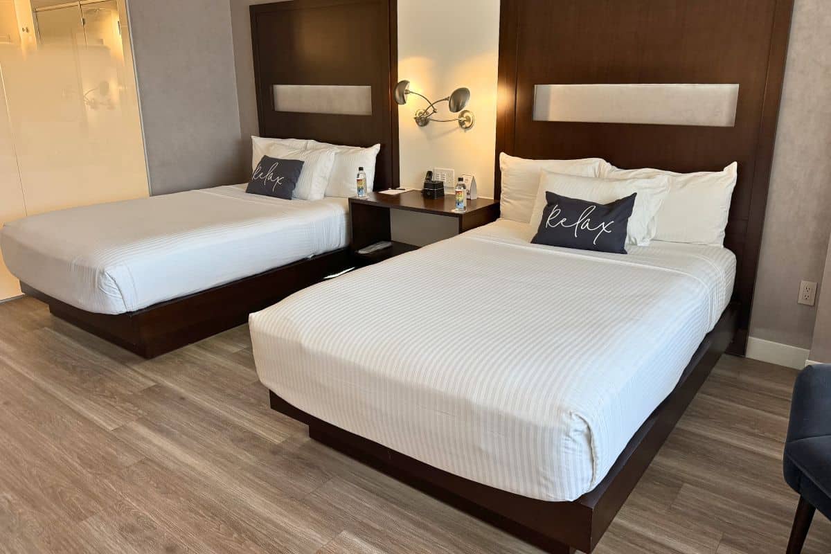Hotel room with two double beds, white linens, "Relax" pillows, wall-mounted lamps, a shared nightstand with water bottles, and wooden floors.