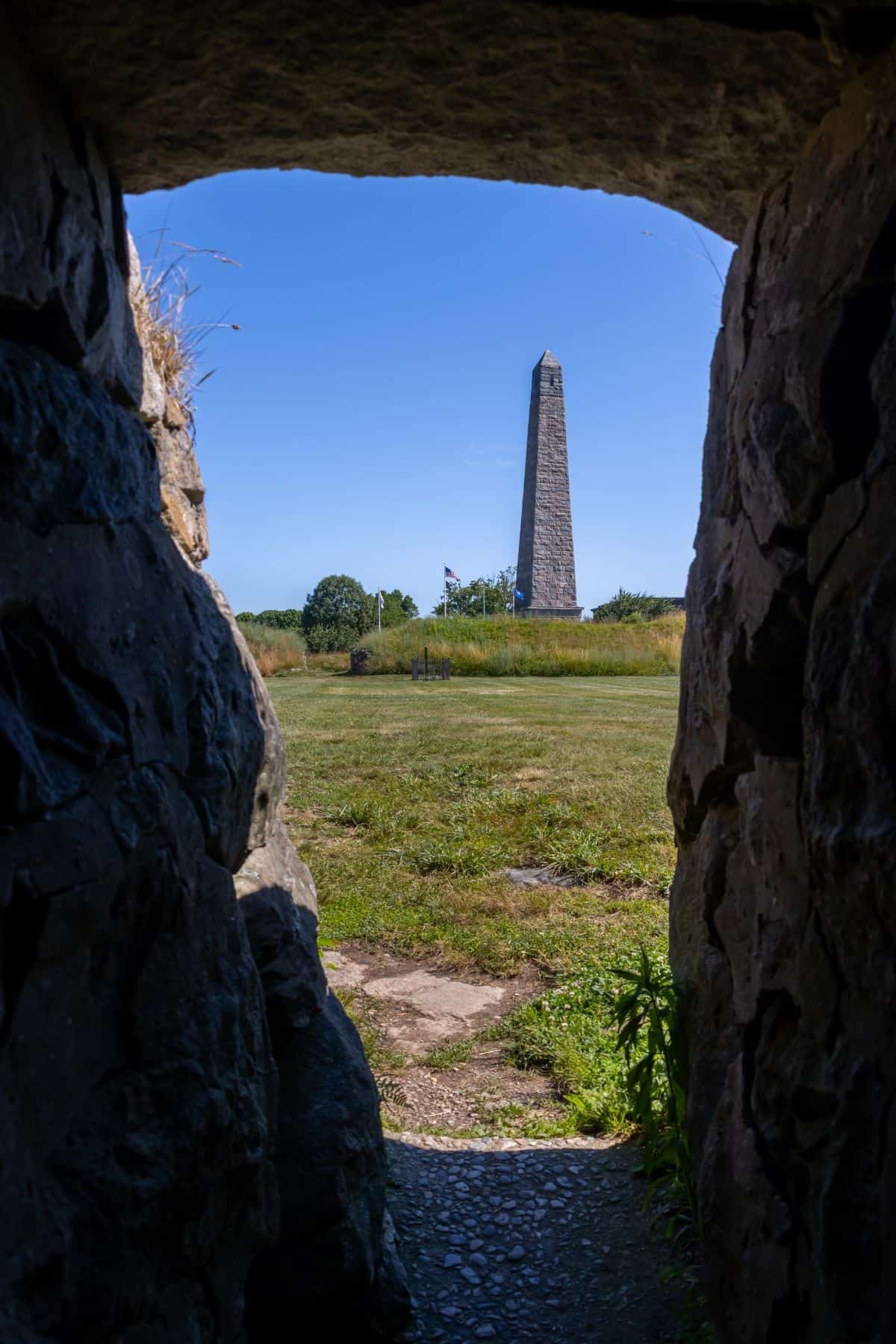 Stone monument stands in an open grassy field, viewed through a rocky tunnel or archway on a clear sunny day.