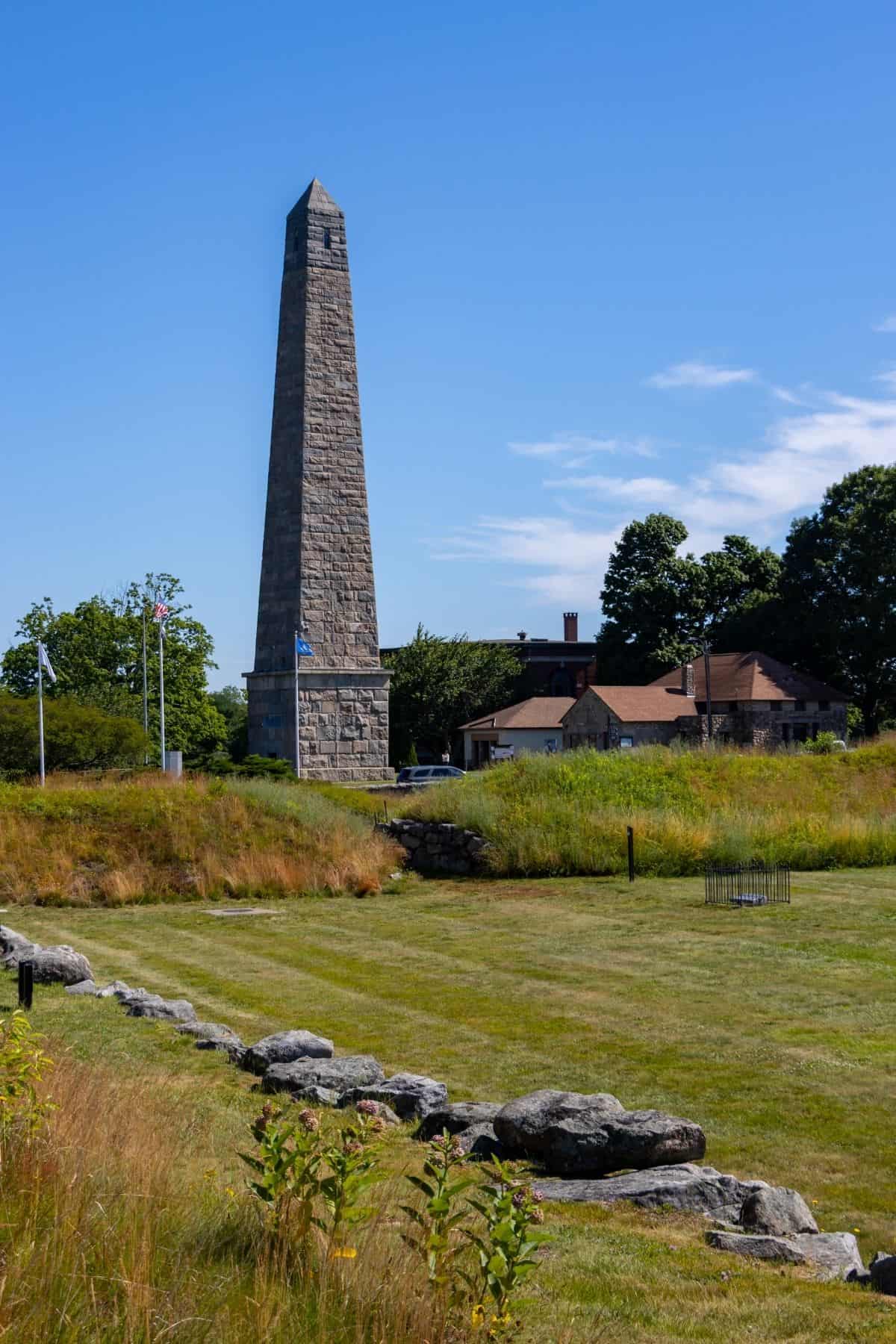 A tall stone obelisk rises beside a historic brick building, surrounded by grass, trees, and a few flags on a clear, sunny day.