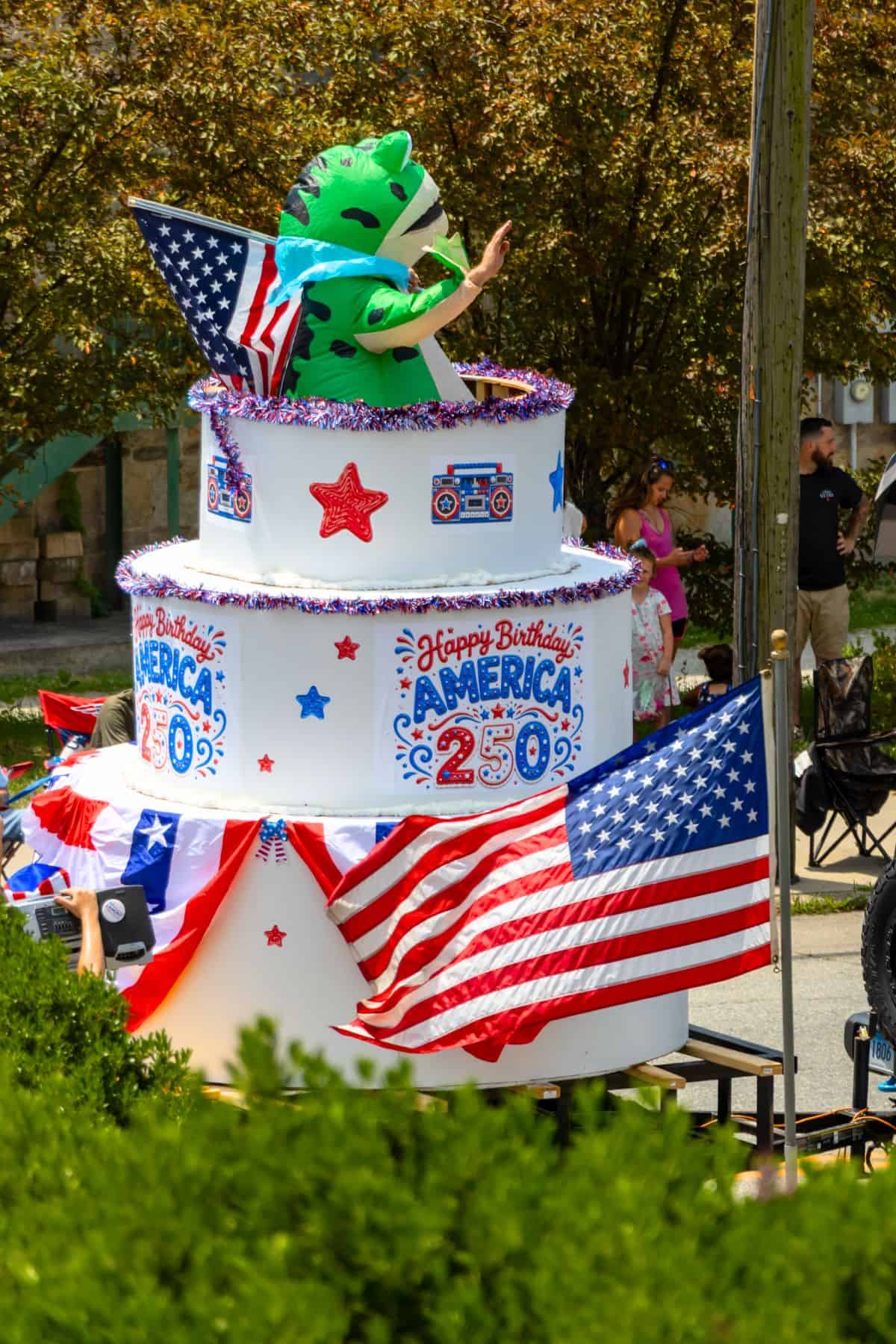 A person in a green dinosaur costume stands inside a large, decorated birthday cake float celebrating America's 250th birthday, with U.S. flags and festive decorations visible.