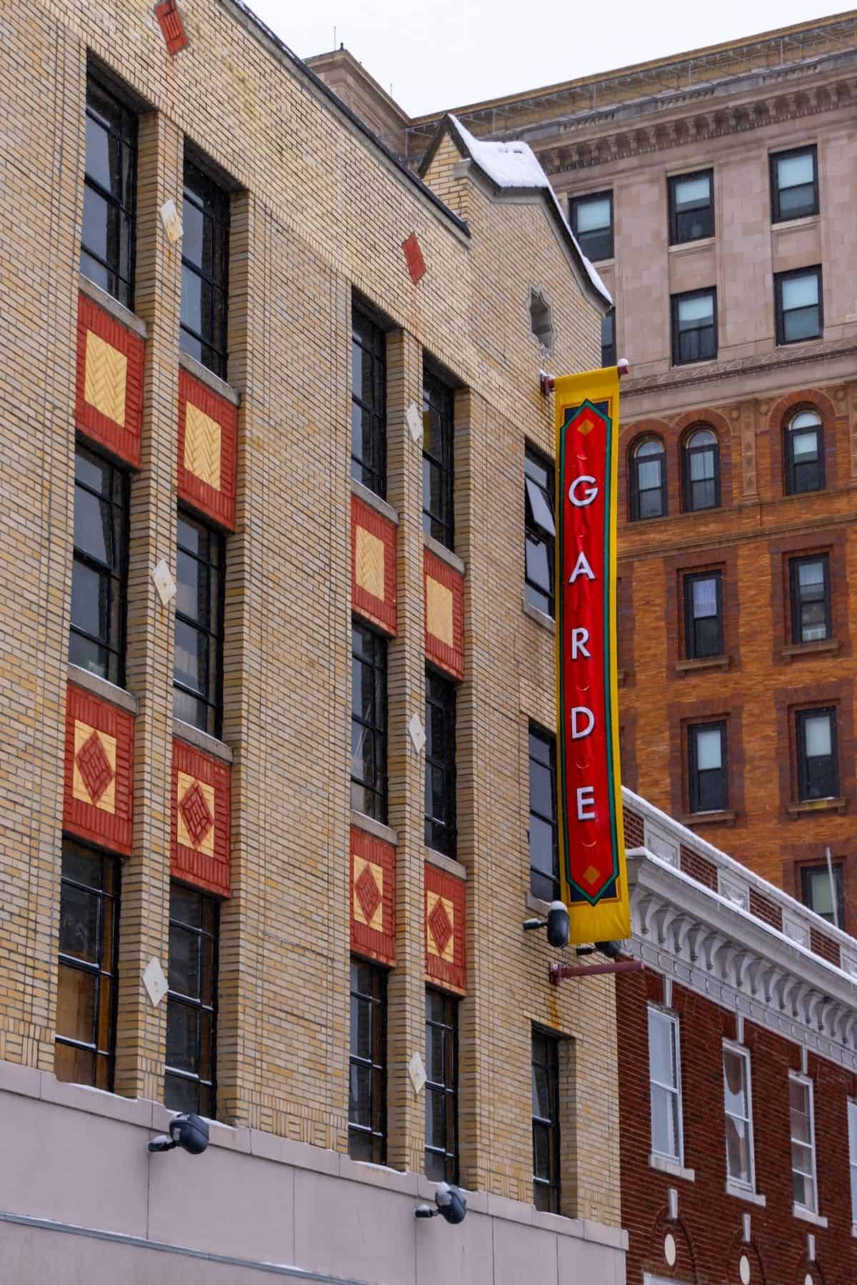 A building with yellow brick facade and red decorative panels features a vertical sign reading "GARDE" in large letters. Another building is visible in the background.