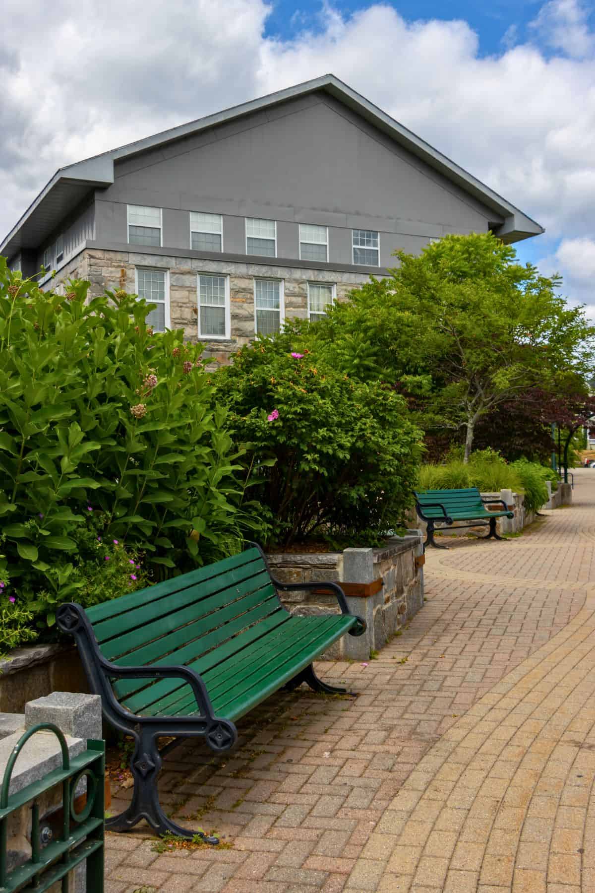 Green benches line a paved walkway in front of a gray and stone building, with bushes and trees alongside under a partly cloudy sky.