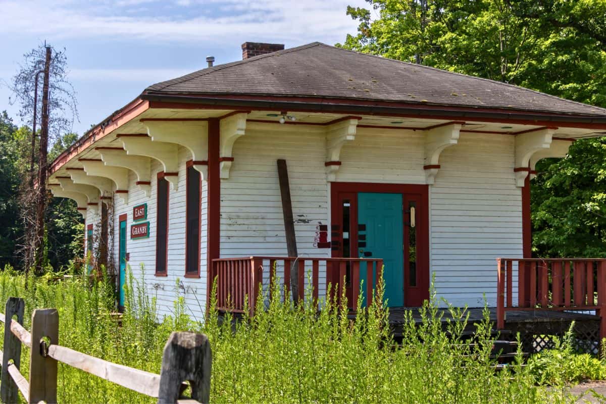 An old, small white building with a green door sits amid overgrown weeds, with a wooden fence and trees in the background under a partly cloudy sky.