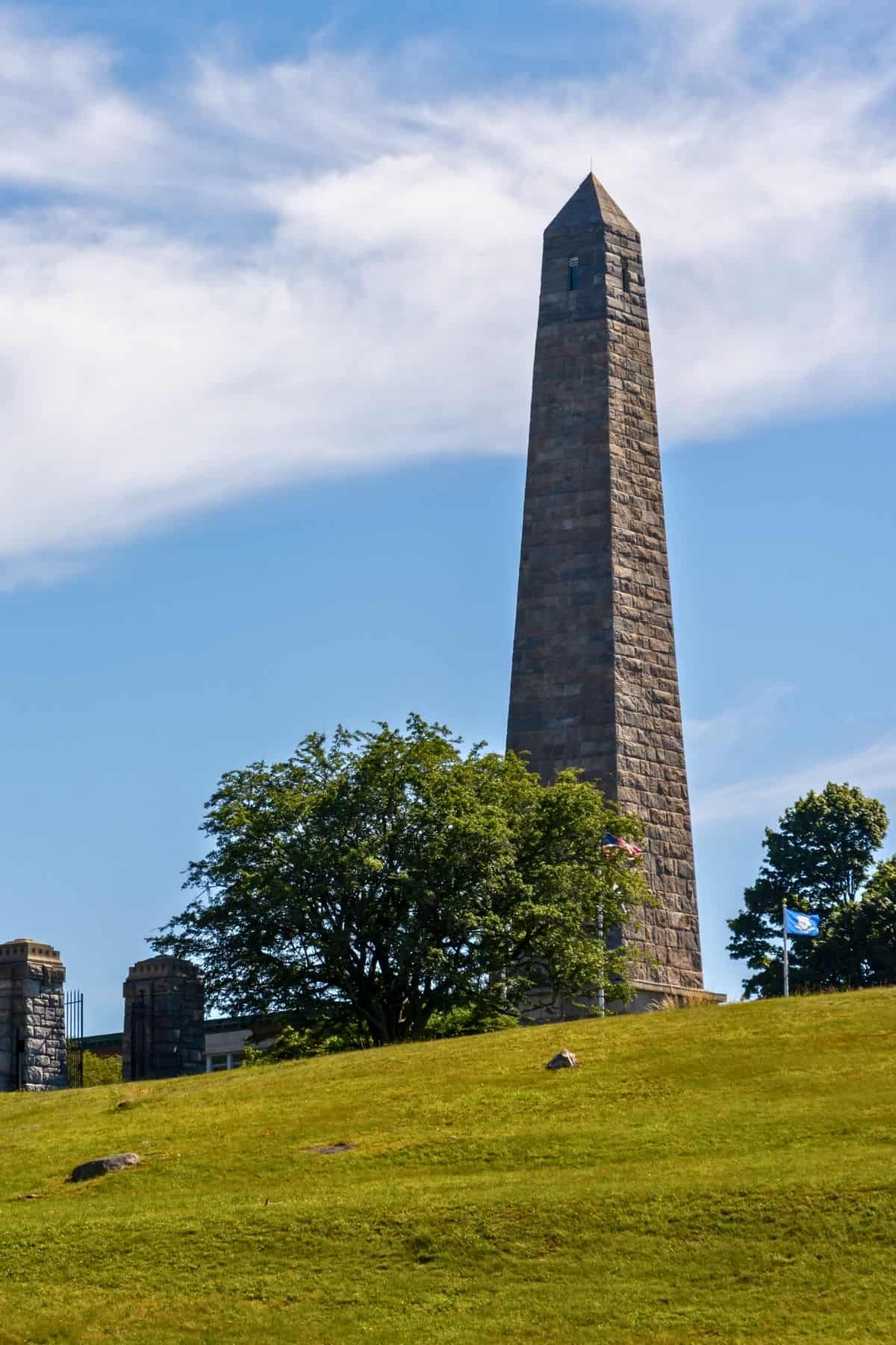 A tall stone obelisk stands atop a grassy hill, with trees and a partly cloudy blue sky in the background.