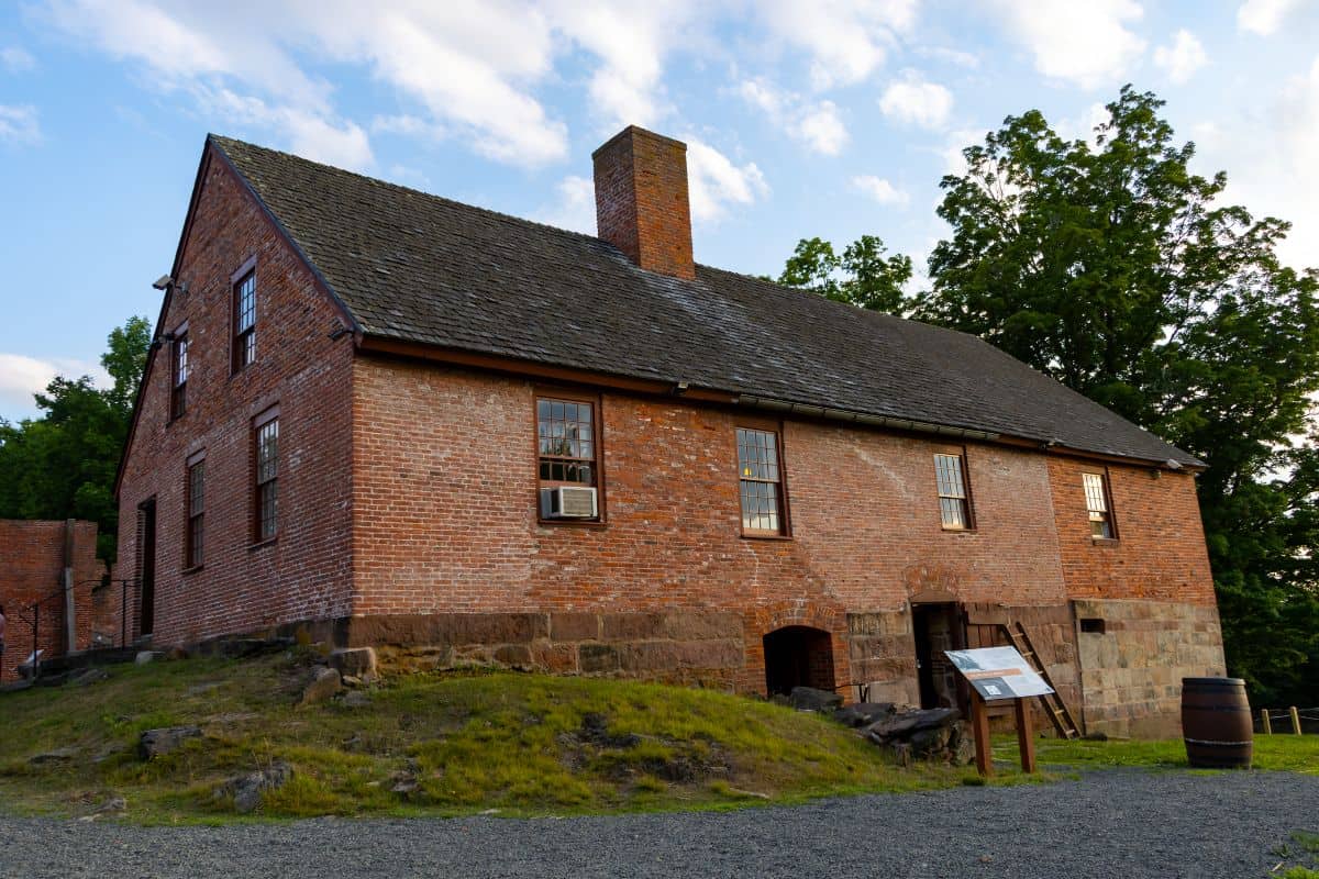 A historic brick building with multiple windows, a shingle roof, chimney, and information signs out front, surrounded by grass and trees.