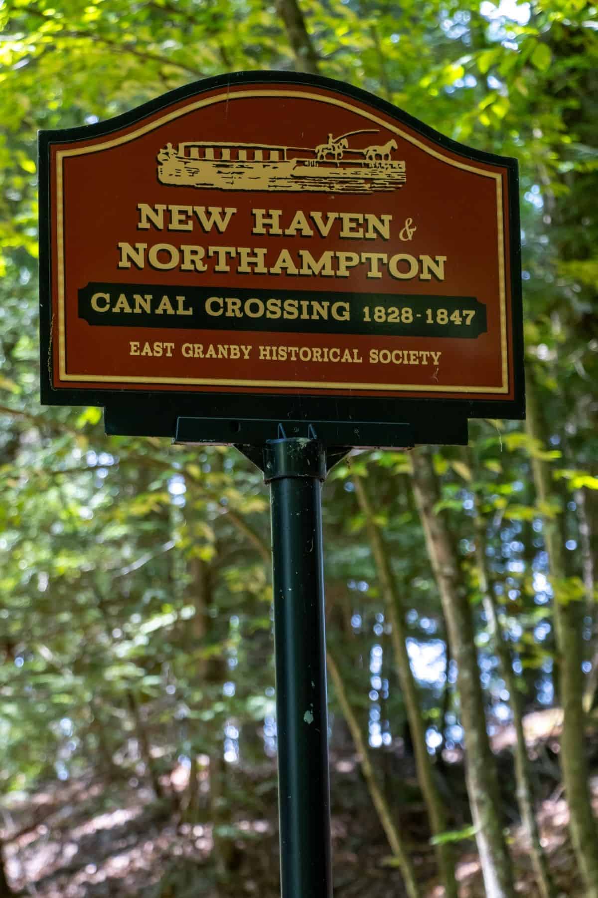 Red and black sign reads "New Haven & Northampton Canal Crossing 1828-1847," with train illustration; trees and greenery in background.
