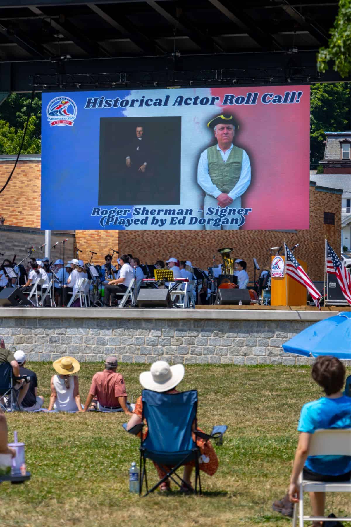 Large outdoor screen displaying "Historical Actor Roll Call!" featuring Roger Sherman, Signer (Played by Ed Dorgan), with people seated on lawn chairs watching a stage below.