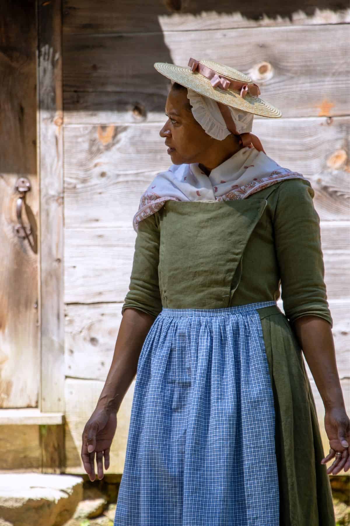 A woman in historical clothing, including a bonnet, green dress, and blue checkered apron, stands outside by a wooden building, looking to her left.