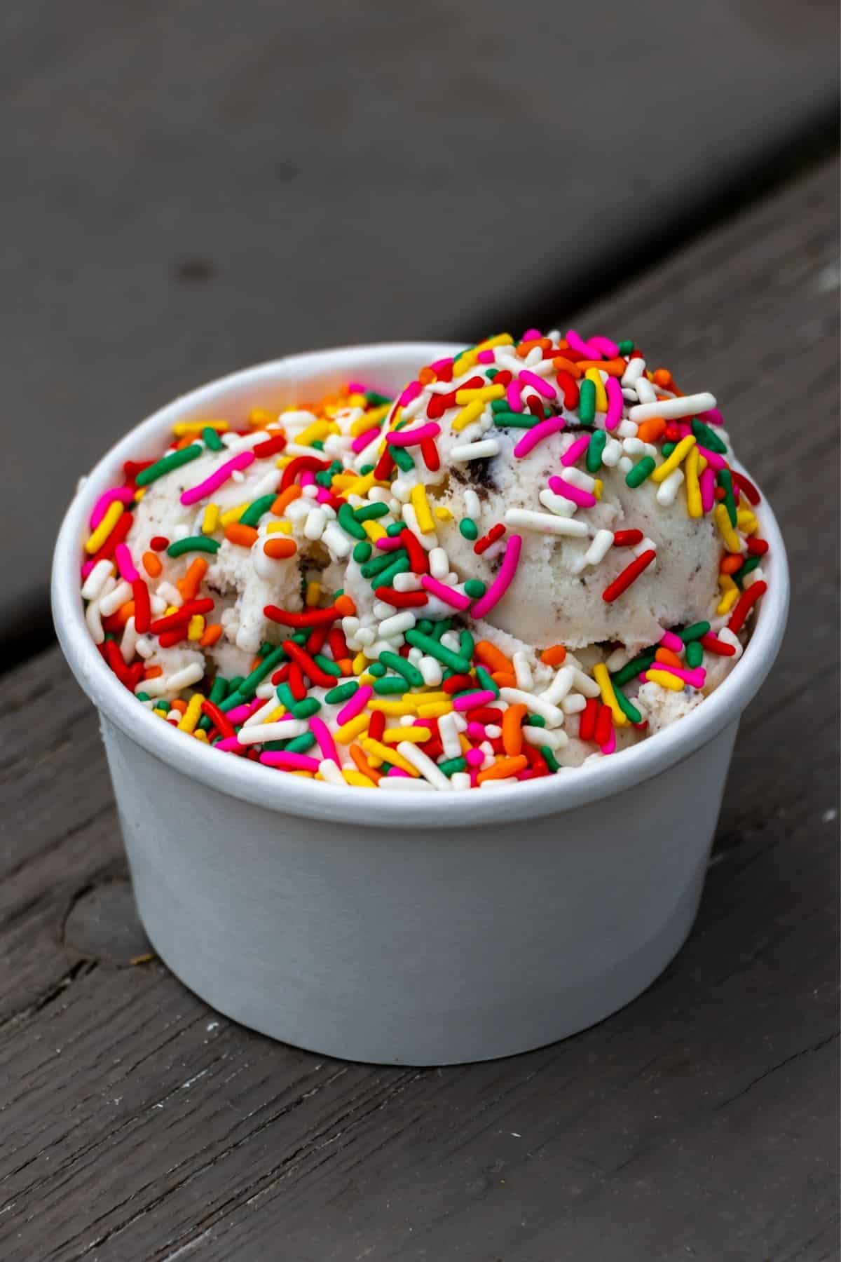 A white paper cup filled with two scoops of ice cream topped with colorful sprinkles, placed on a wooden surface.