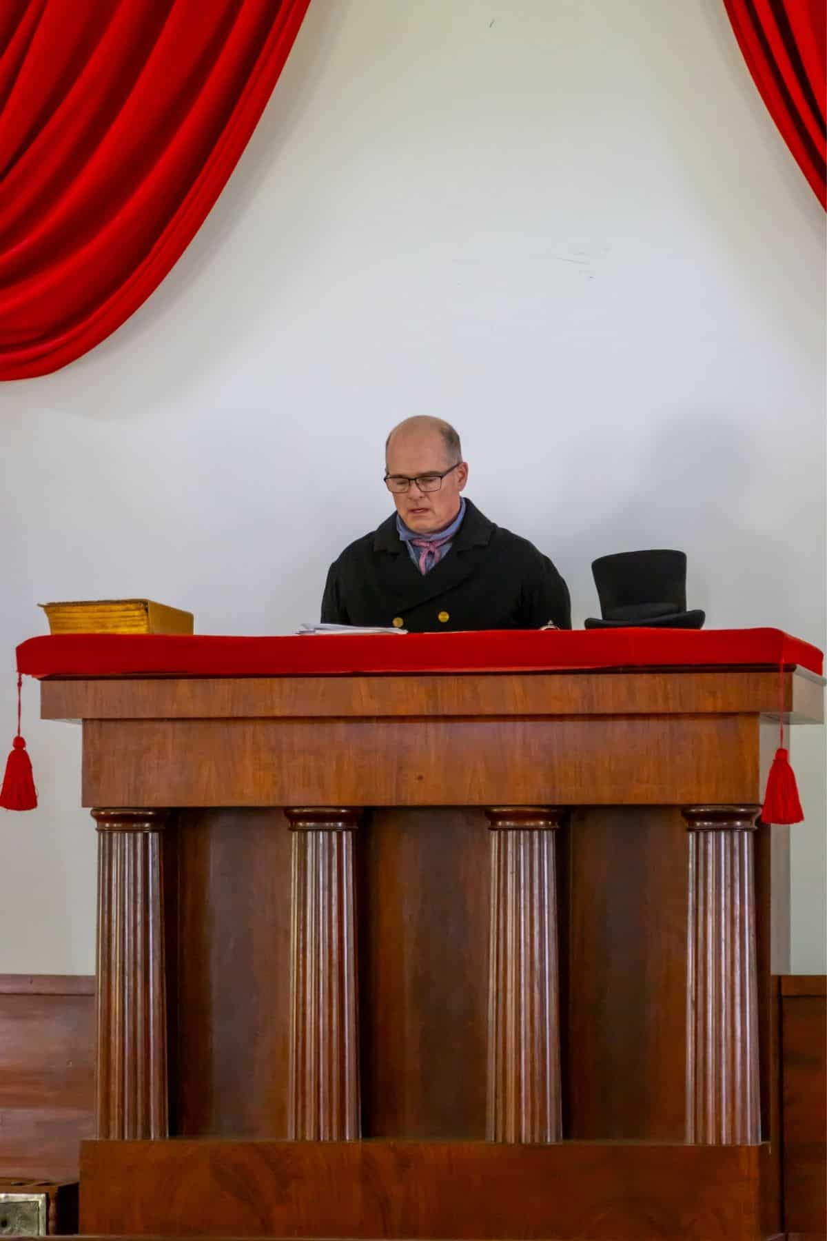 A man in a judge’s robe stands behind a wooden podium with a red cloth, reading papers; a black top hat and a box are on the podium.