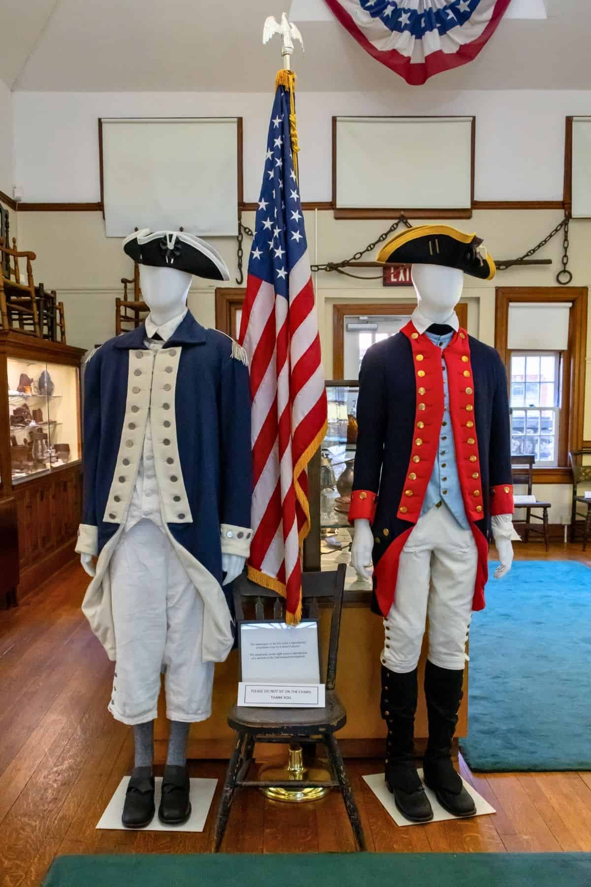 Two mannequins display Revolutionary War-era military uniforms beside an American flag in a museum setting.