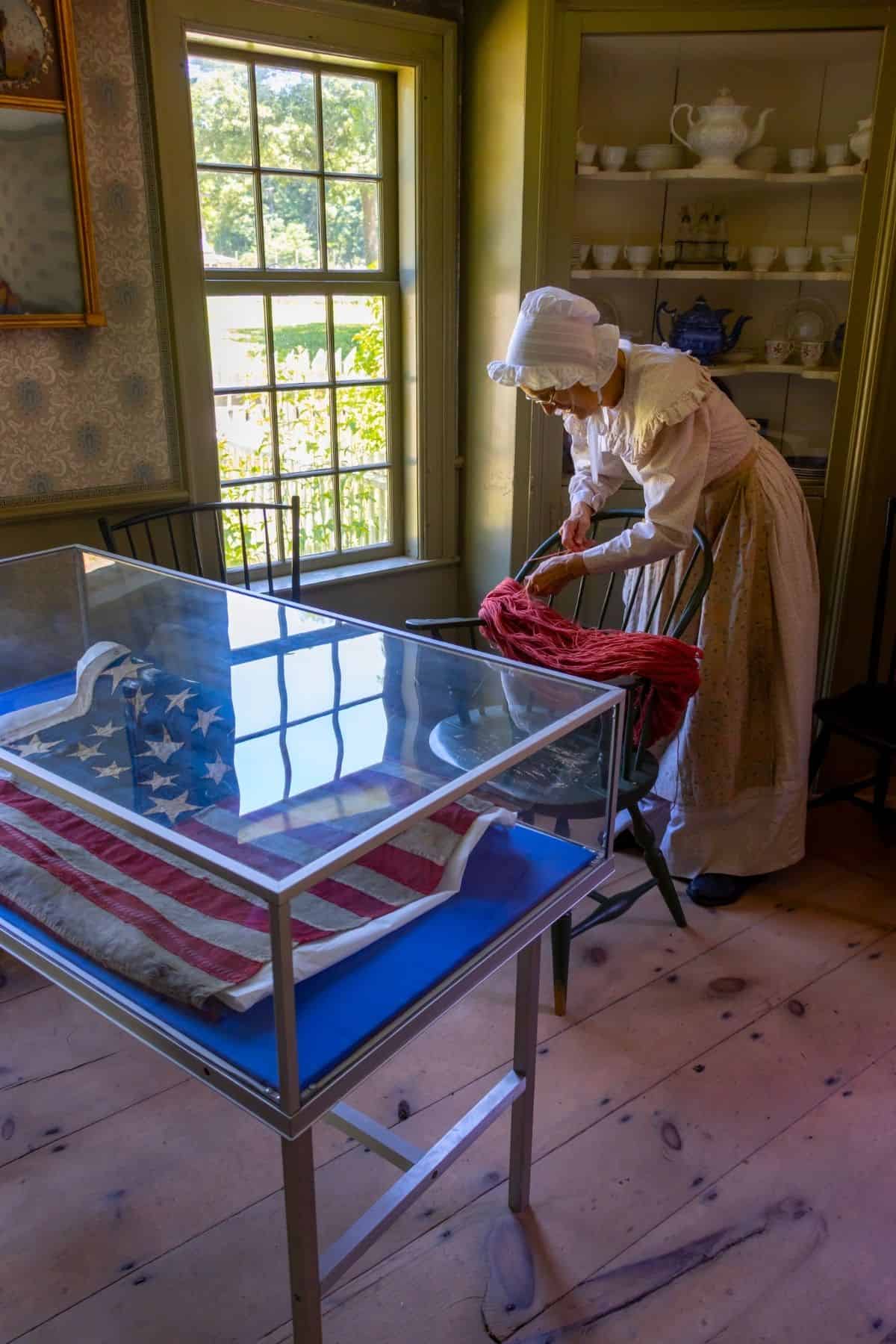 A woman in colonial attire stands near a spinning wheel in a historic room with a glass case displaying an old American flag.