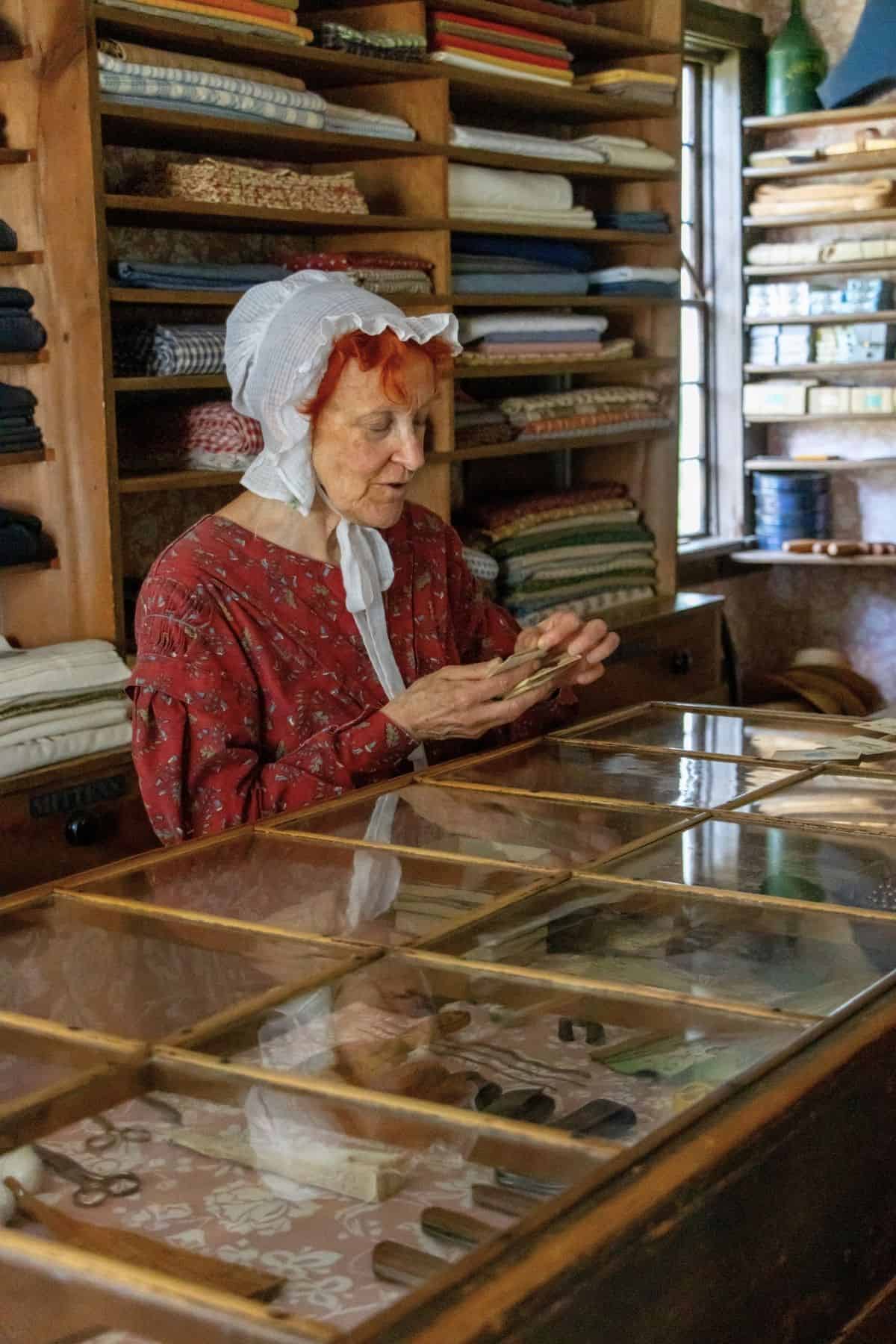 An elderly woman in historical clothing stands behind a glass display counter in a fabric store, handling some money. Shelves with folded fabric are visible in the background.