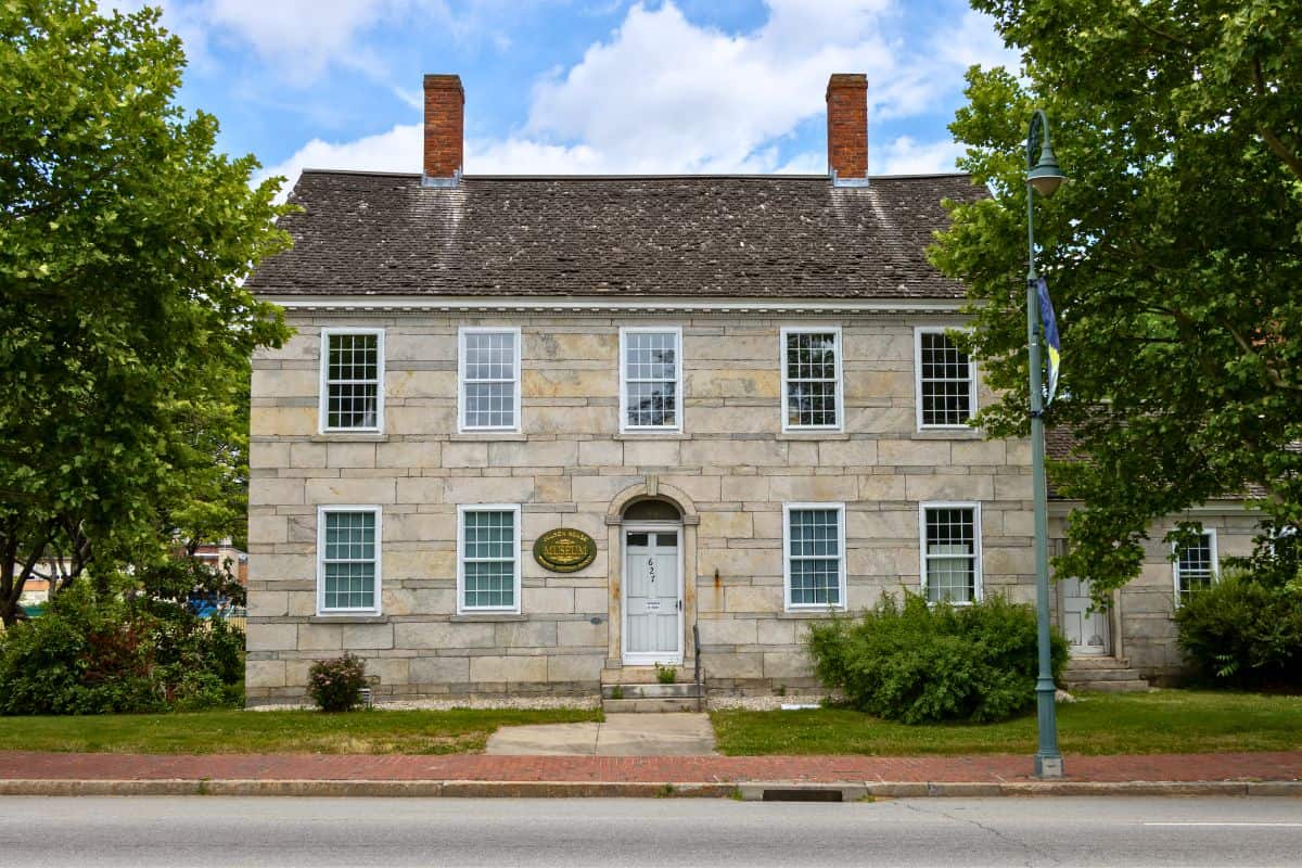 A two-story stone building with a central door, twelve windows, two brick chimneys, and a plaque near the entrance, surrounded by trees and a sidewalk.