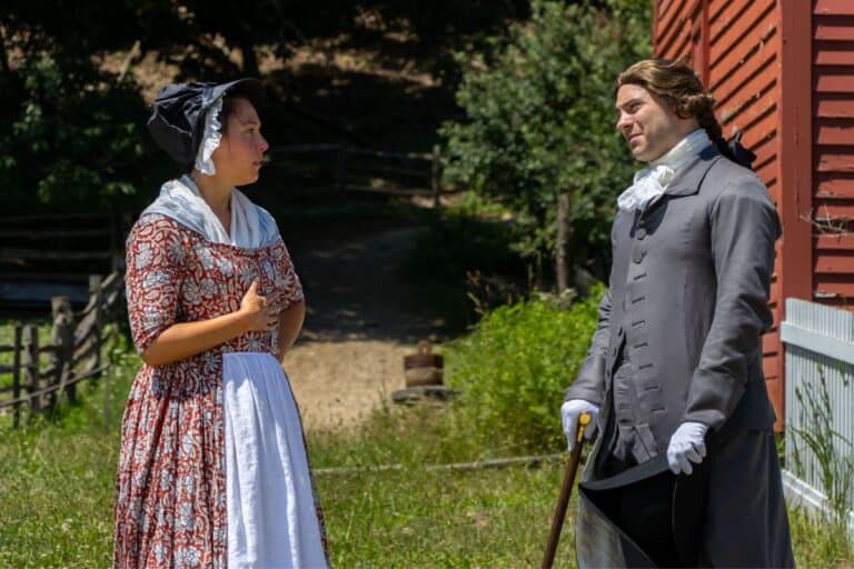 A woman in a patterned dress and bonnet speaks to a man in 18th-century attire with a cane, outside near a red wooden building and greenery.