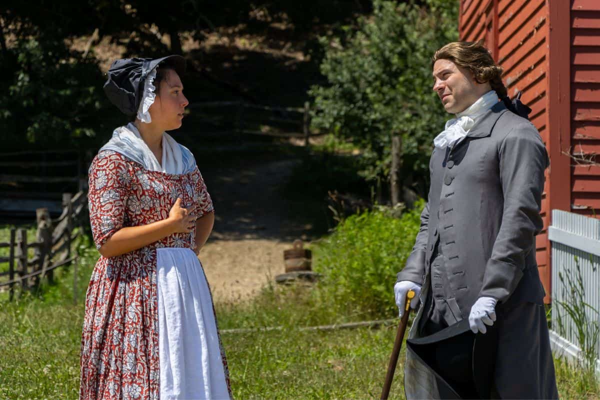 A woman in a patterned dress and bonnet speaks to a man in 18th-century attire with a cane, outside near a red wooden building and greenery.