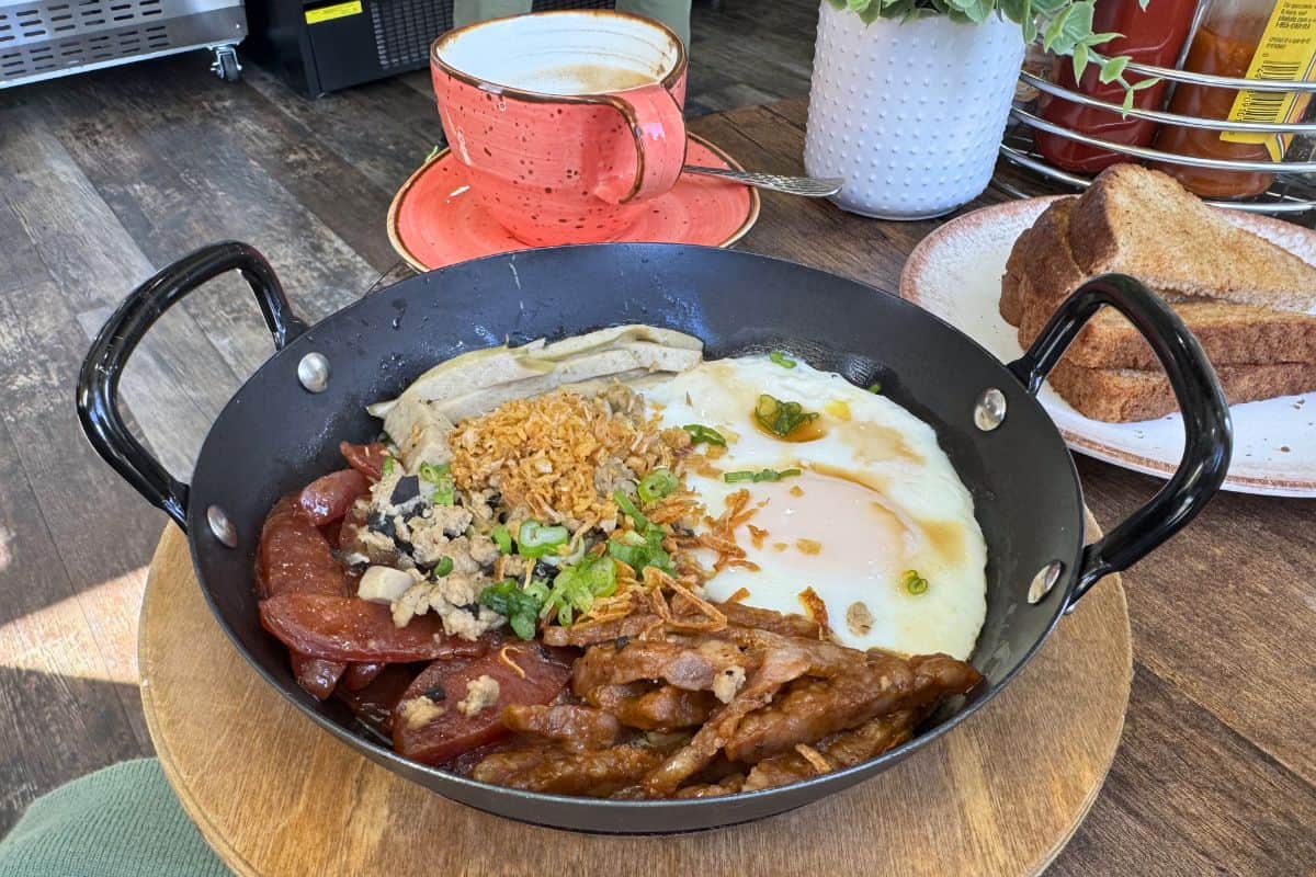 A breakfast skillet with two fried eggs, sliced sausage, shredded meat, chopped green onions, crispy bits, toast on a separate plate, and a cup of coffee on a table.