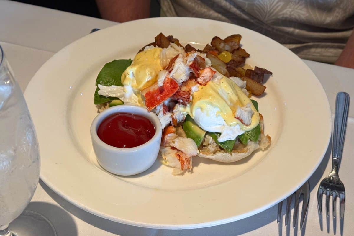 Plate of eggs benedict with lobster and avocado, served with roasted potatoes and a side of ketchup on a white tablecloth next to a glass of water.