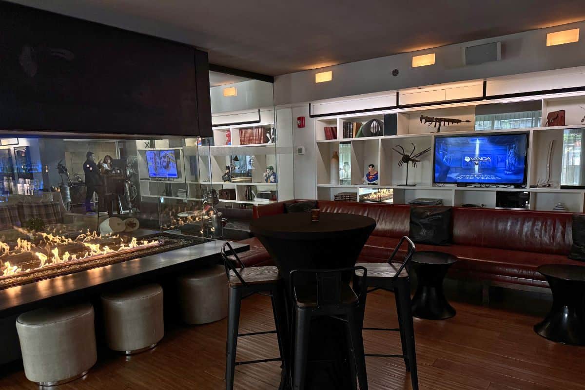 Modern lounge with a glass fireplace, red leather couches, round tables, stools, and wall-mounted TVs. People are reflected in mirrored shelves behind the seating area.