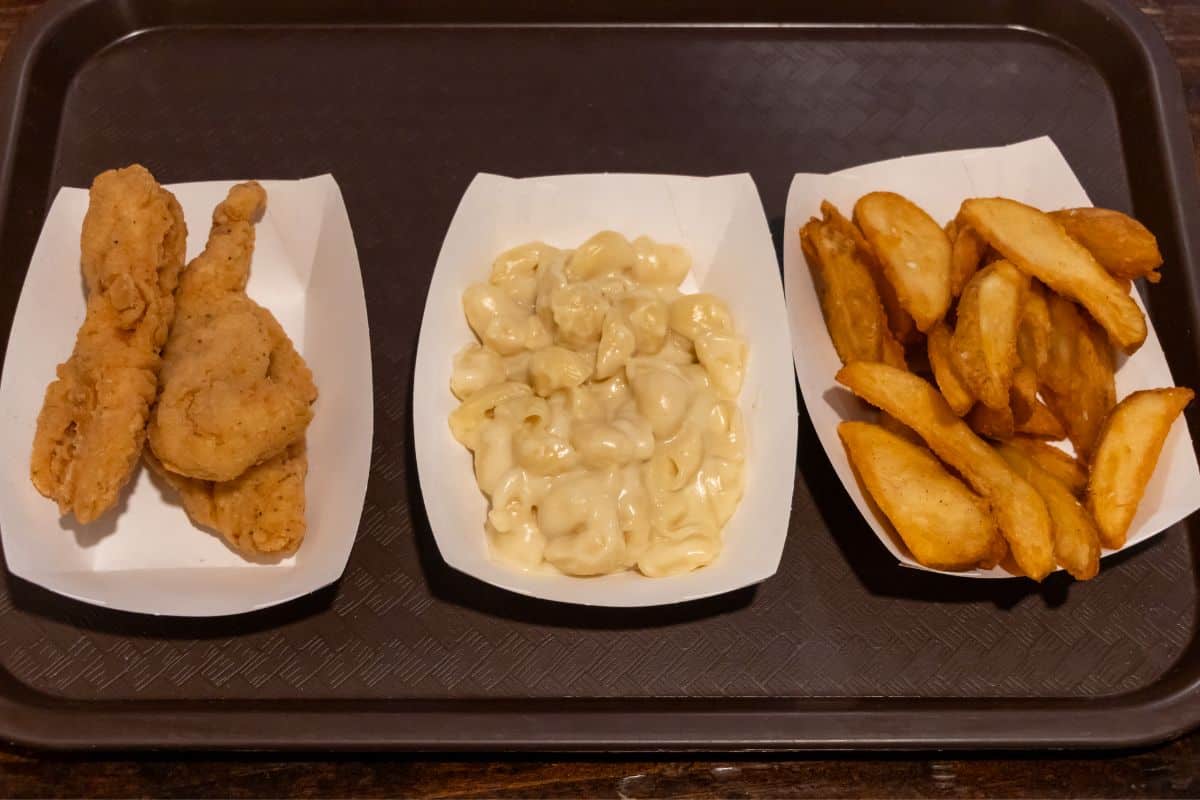 A tray with three paper containers holding fried chicken drumsticks, macaroni and cheese, and potato wedges.