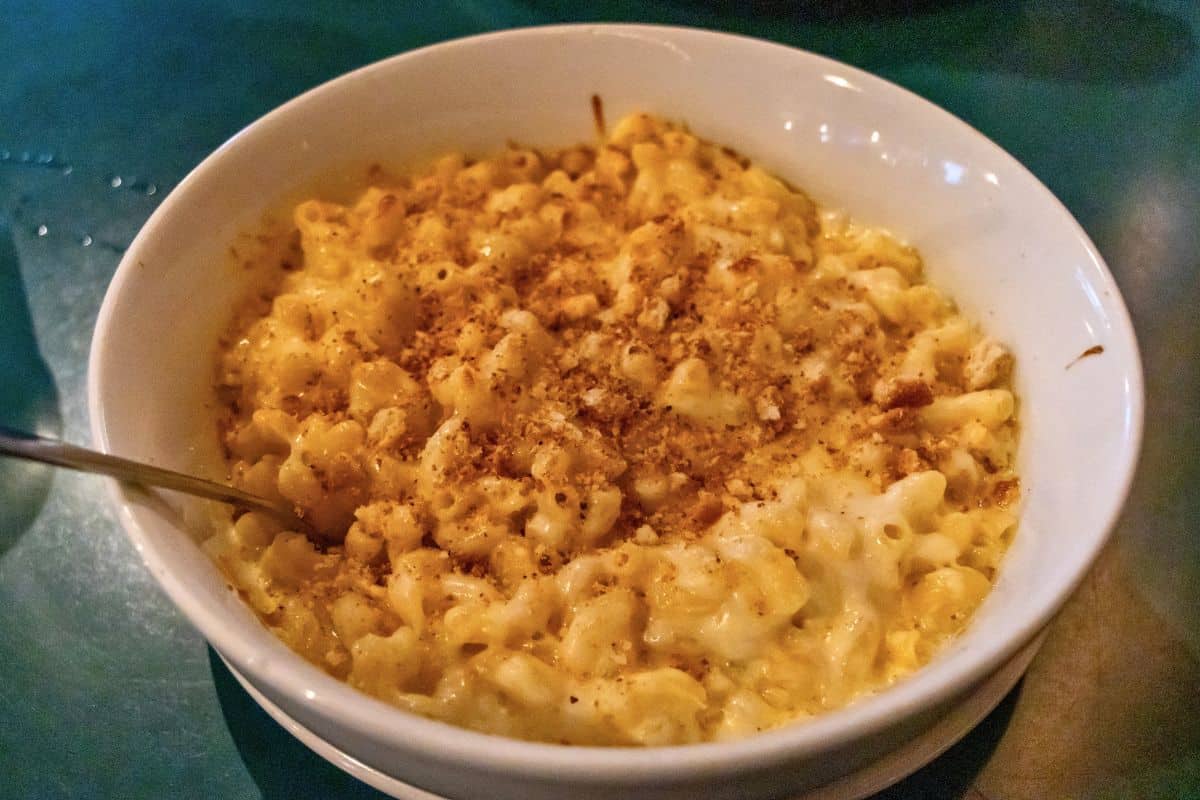 A white bowl filled with macaroni and cheese topped with breadcrumbs, placed on a dark green table, with a spoon on the left side inside the bowl.