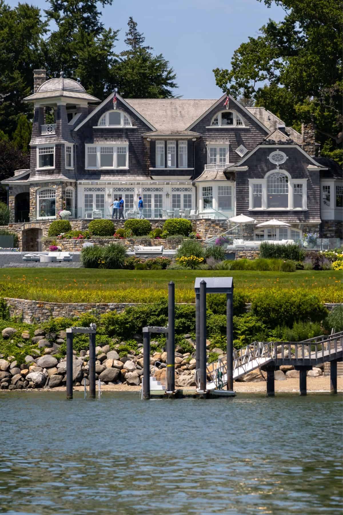 Large, gray-shingled waterfront house with white trim, landscaped garden, and dock, viewed from the water on a sunny day.