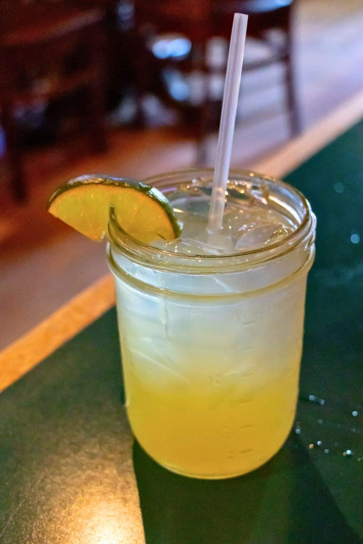 A mason jar filled with a yellow beverage, ice cubes, a lime wedge garnish, and a clear straw sits on a green table.