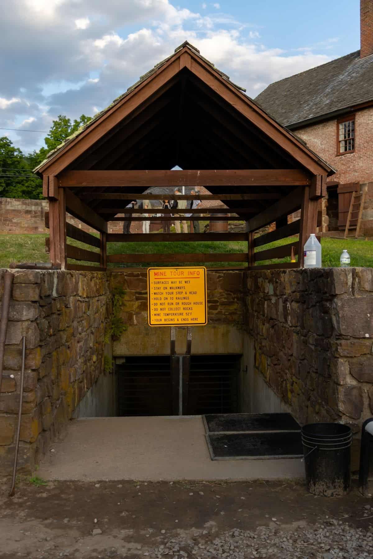 A covered stone entrance to a mine with a yellow information sign about mine tours, flanked by bottles and a bucket, with buildings and grass in the background.