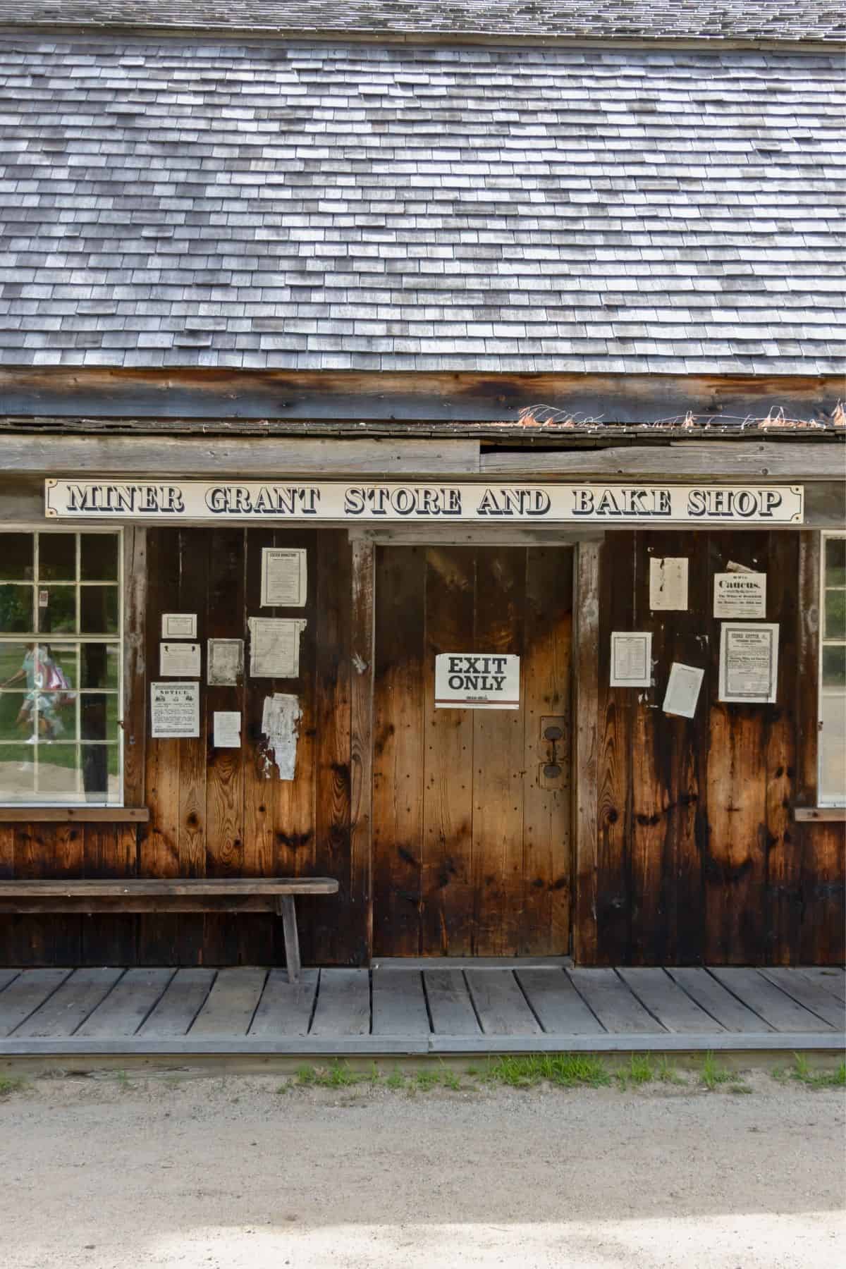 Wooden storefront reading “Miner Grant Store and Bake Shop,” with posted notices, a bench, and an “Exit Only” sign on the door.