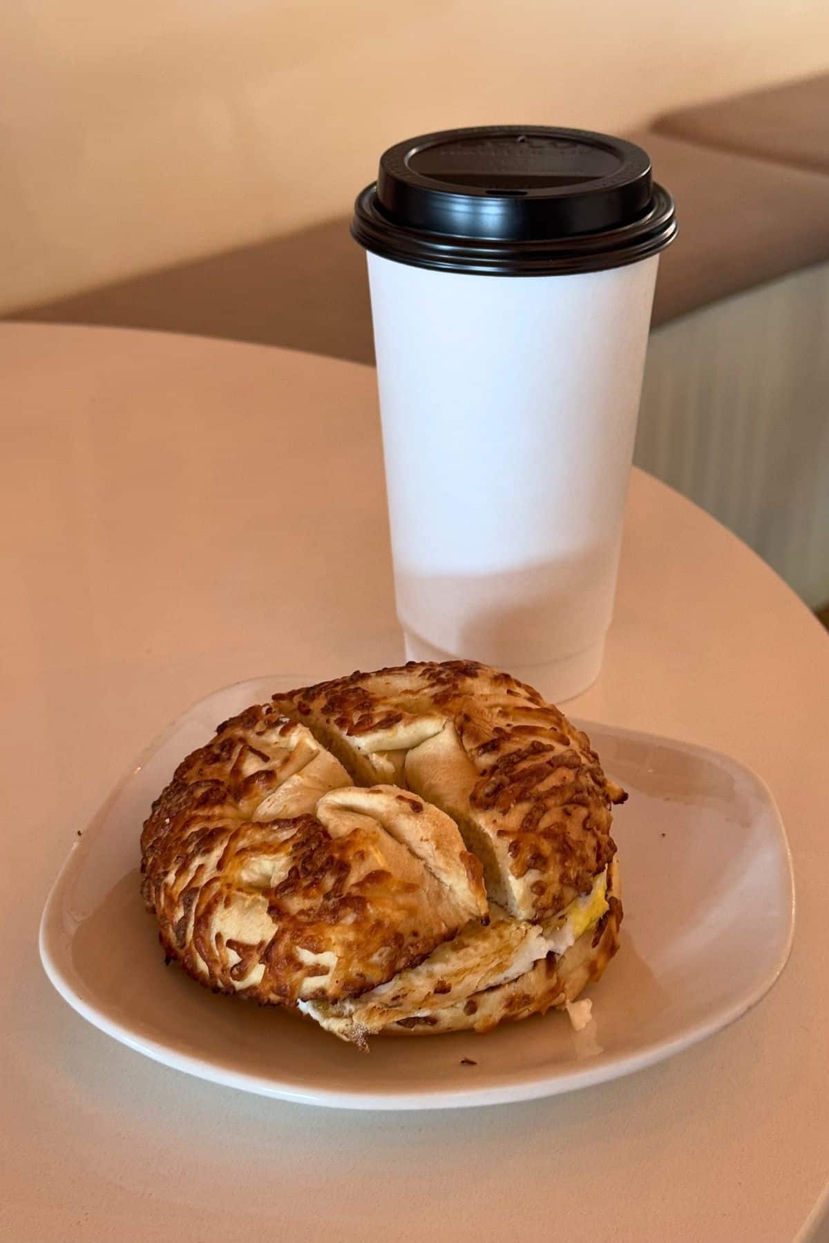 A paper cup with a black lid and a cheese-topped bagel sandwich on a white plate, placed on a light-colored table.