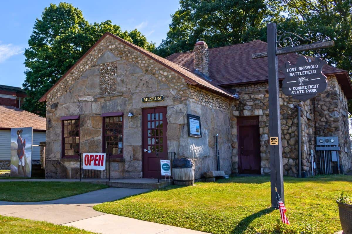 Stone museum building with a red door and "OPEN" sign, located at Fort Griswold Battlefield, Connecticut State Park, on a sunny day with green grass and trees in the background.