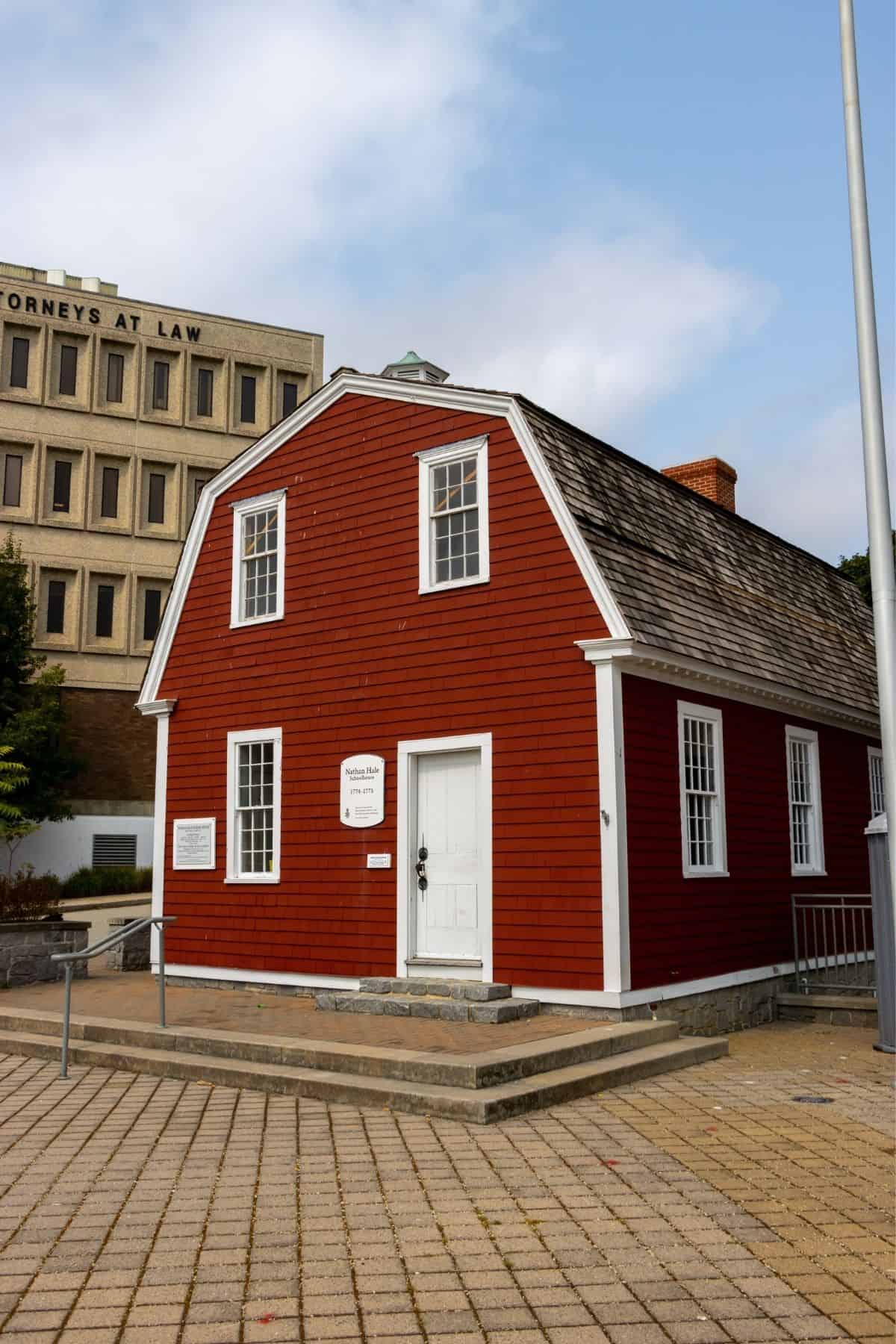 A small, red, two-story historic building with white trim stands on a brick plaza, with a modern office building visible in the background.
