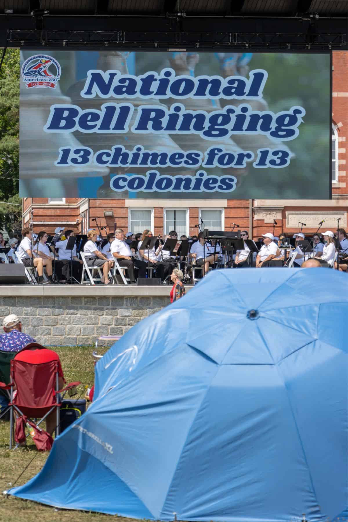 Large outdoor screen displays "National Bell Ringing, 13 chimes for 13 colonies" with a band performing on stage and a blue tent in the foreground.