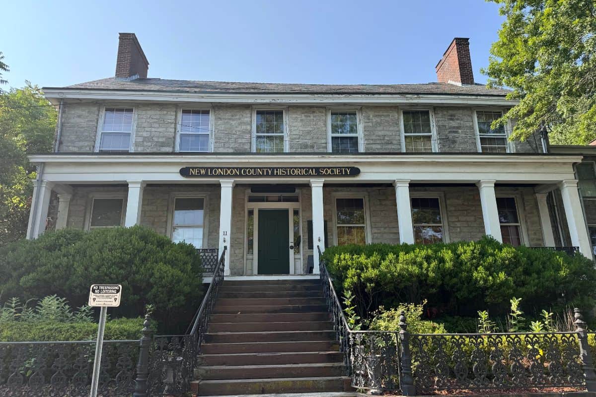 A two-story stone building with a sign reading "New London County Historical Society" above the entrance, surrounded by greenery and an iron fence.