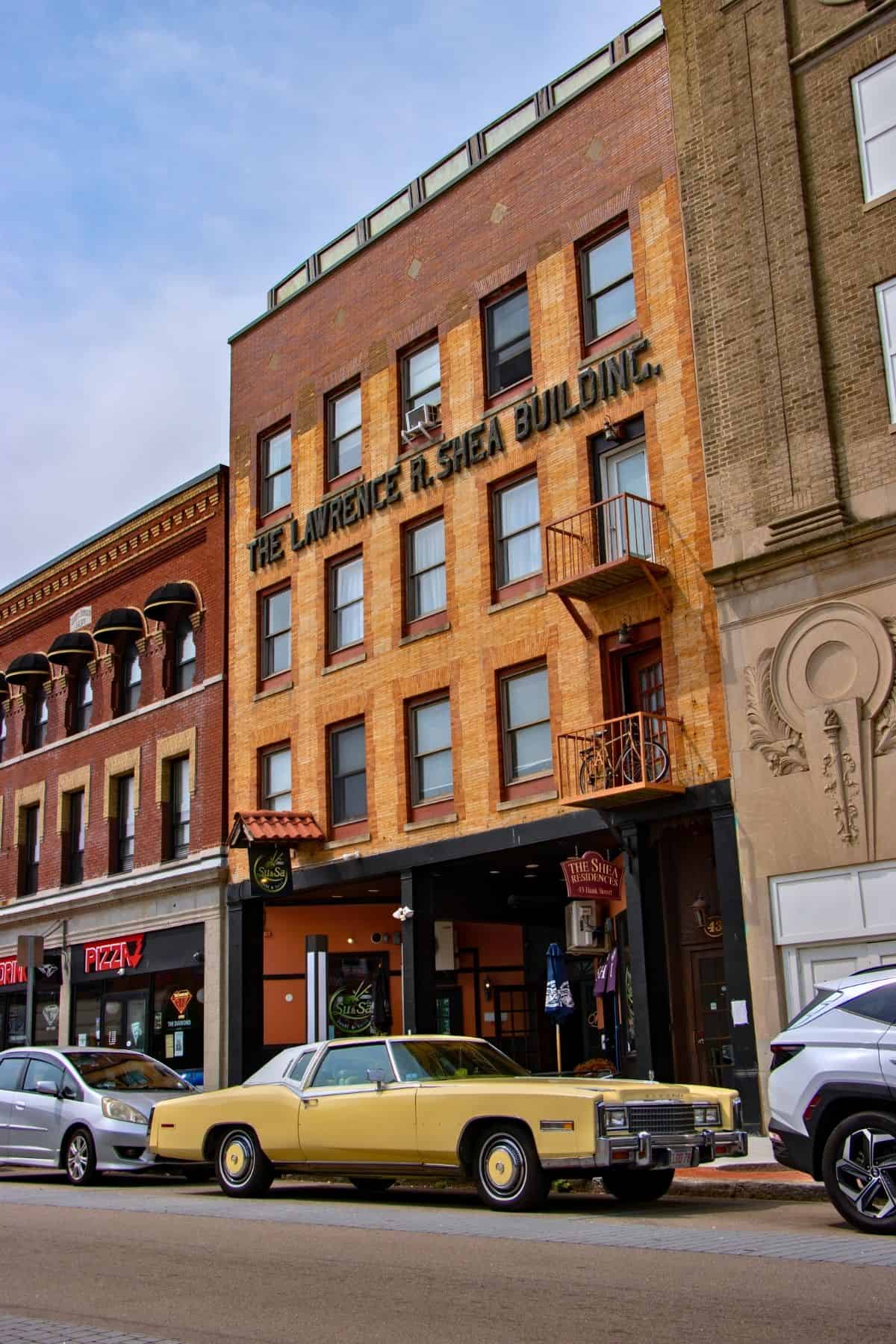 A vintage yellow car is parked on the street in front of the Lawrence R. Shea Building, a multi-story brick structure with shops at ground level.