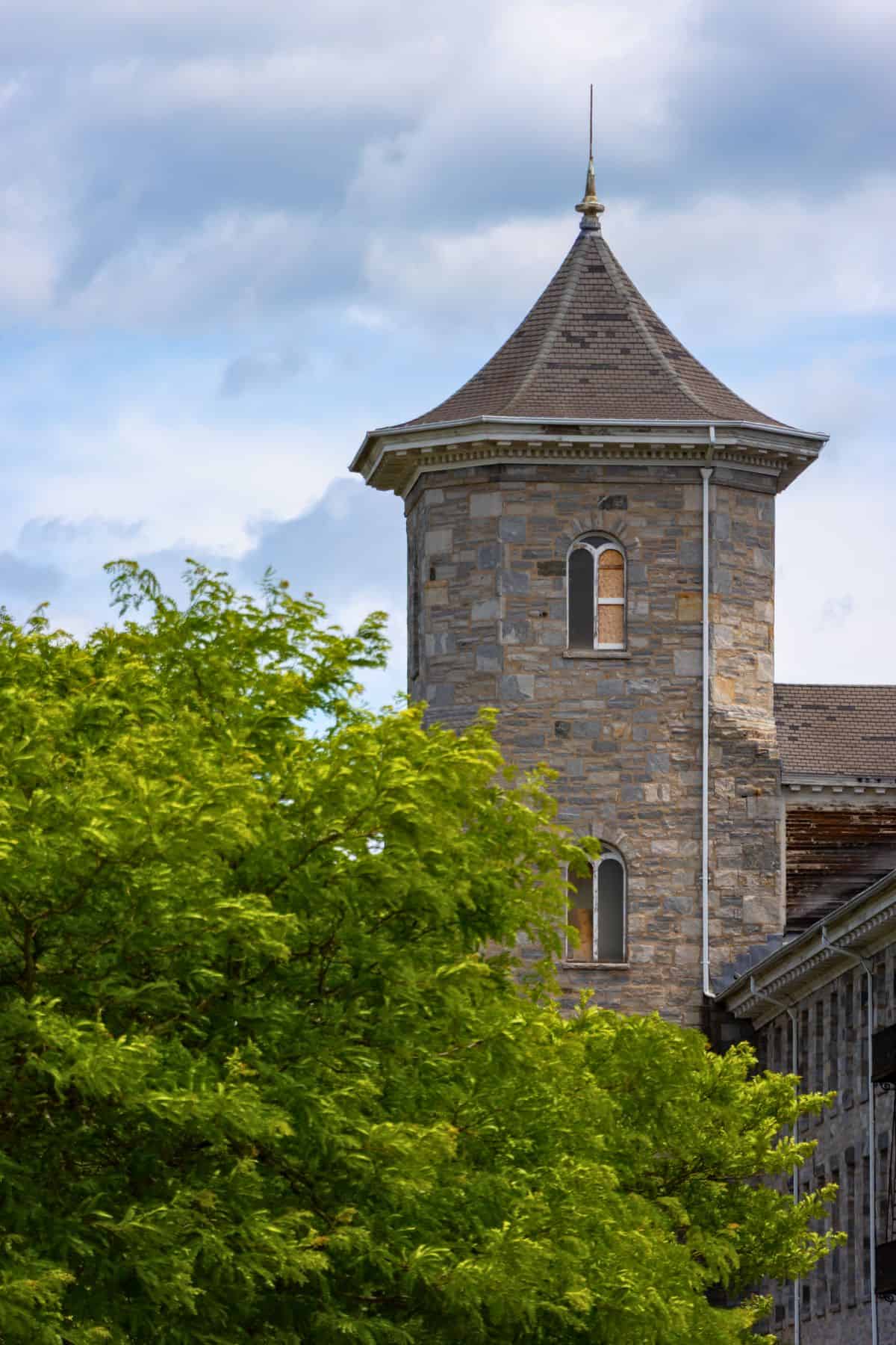 Stone tower with arched windows and a conical roof stands behind leafy green trees under a partly cloudy sky.