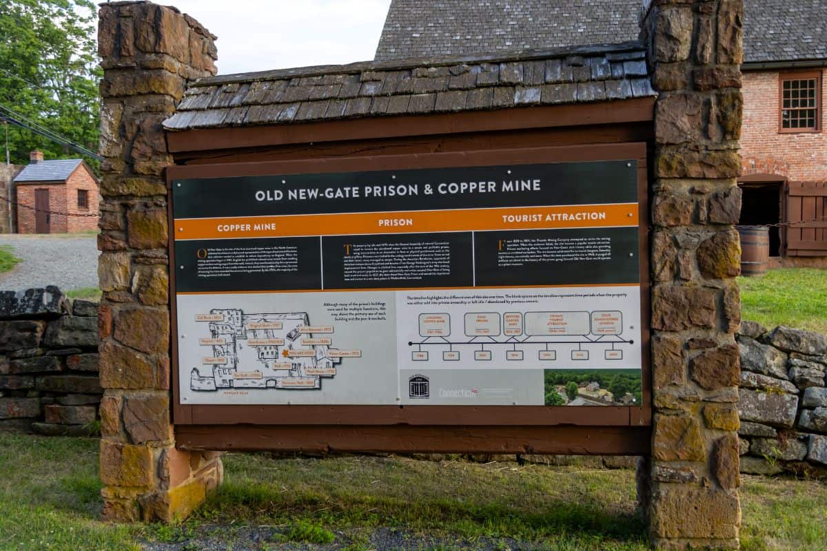 Informational sign for Old New-Gate Prison & Copper Mine, detailing its history as a mine, prison, and tourist site, with diagrams and a timeline, set between two stone pillars.
