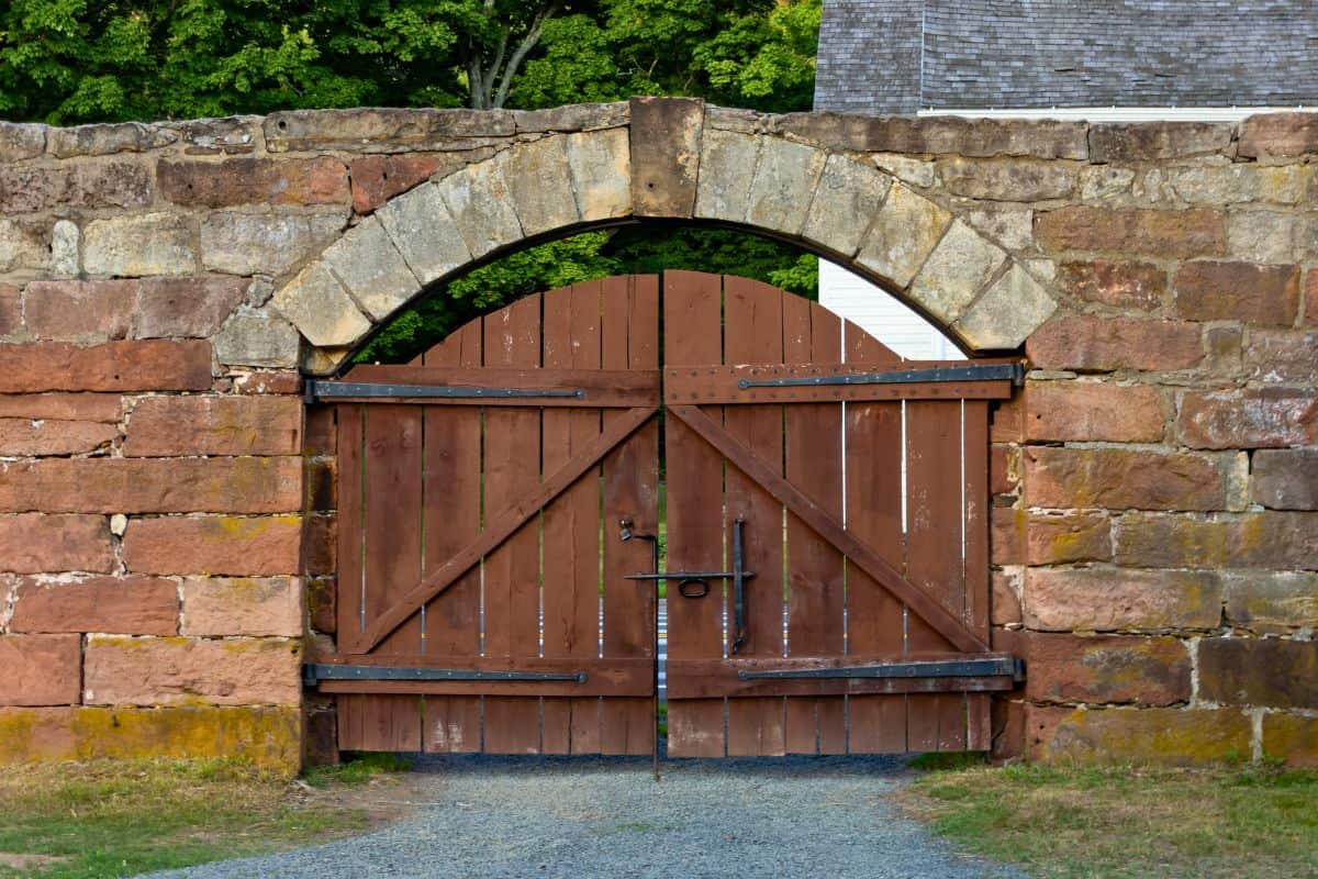 Wooden double gate set in a stone archway, with metal hinges and latch, stands closed on a gravel path. Green trees and part of a building roof are visible in the background.