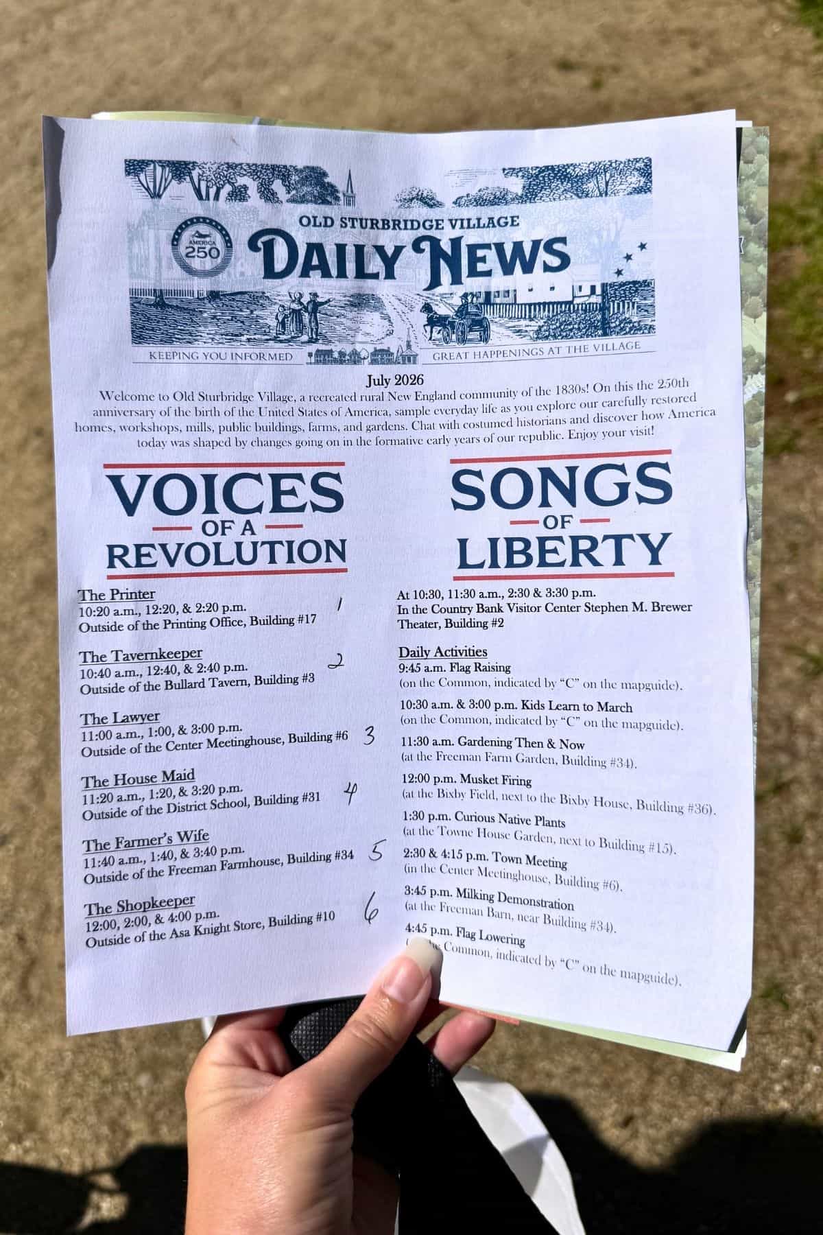 A person holds a copy of Old Sturbridge Village’s Daily News listing July 2026 events, including “Voices of a Revolution” and “Songs of Liberty” with dates, times, and locations.