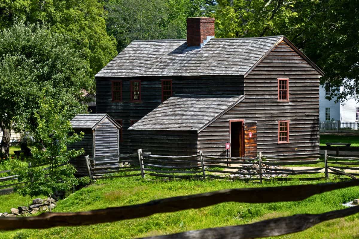 A wooden colonial-style house with red-trim windows sits behind a split-rail fence, surrounded by green grass and trees on a sunny day.