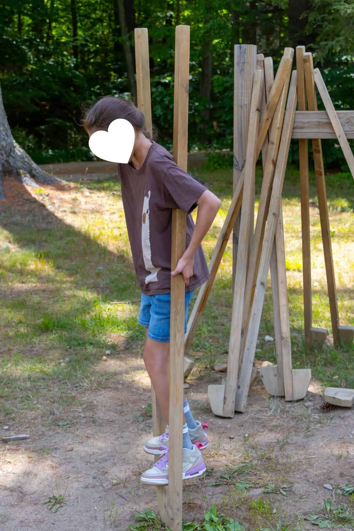 A child in shorts and a brown shirt walks on wooden stilts outdoors, with more stilts leaning against a wooden frame nearby.