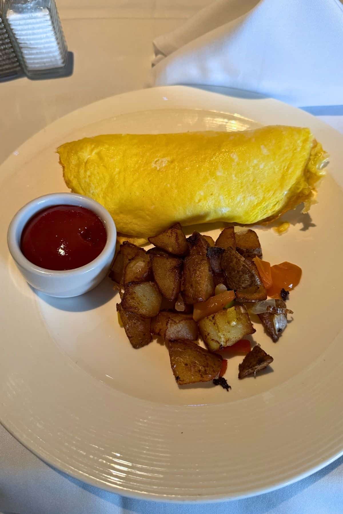 A yellow omelette served with a side of diced roasted potatoes and a small cup of ketchup on a white plate.