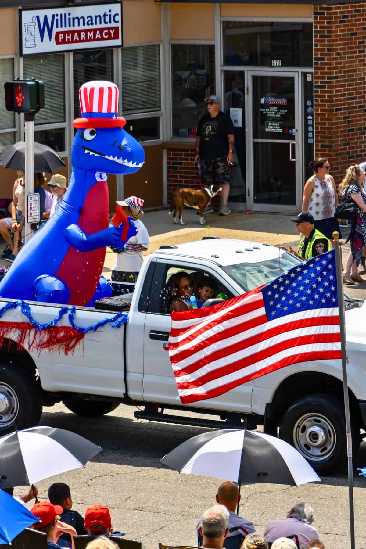 A white pickup truck with an American flag and a large inflatable dinosaur in a red, white, and blue hat drives in a parade; spectators watch from the sidewalk.